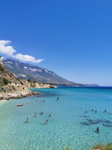 Scenic coastline with clear blue water, swimmers, and mountains under a bright summer sky.